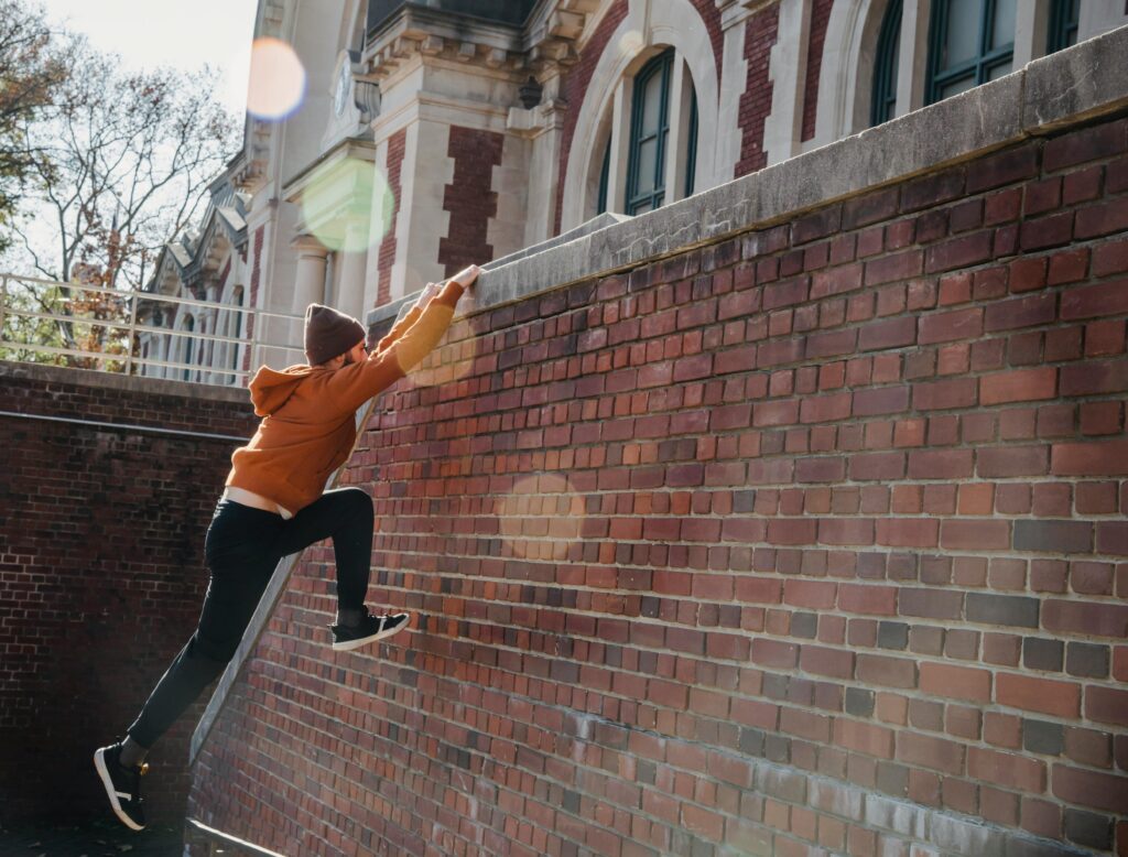Side view full body of determined male in hat jumping on wall and grabbing onto railing during training on street